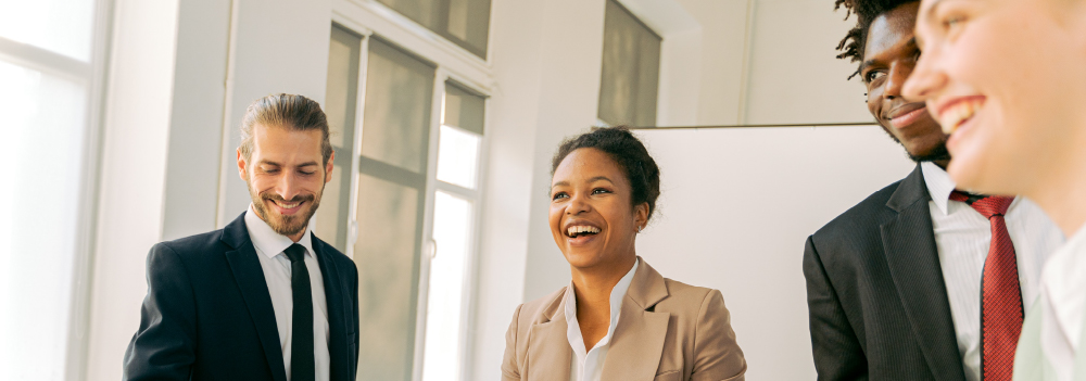 Four coworkers, two men and two women, smile and talk while standing around a conference table.