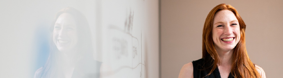 Smiling woman with long red hair stands near a whiteboard in an office conference room.