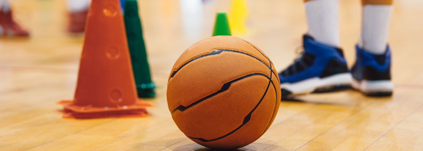 Basketball on gym floor next to an orange cone and a player wearing tennis shoes.